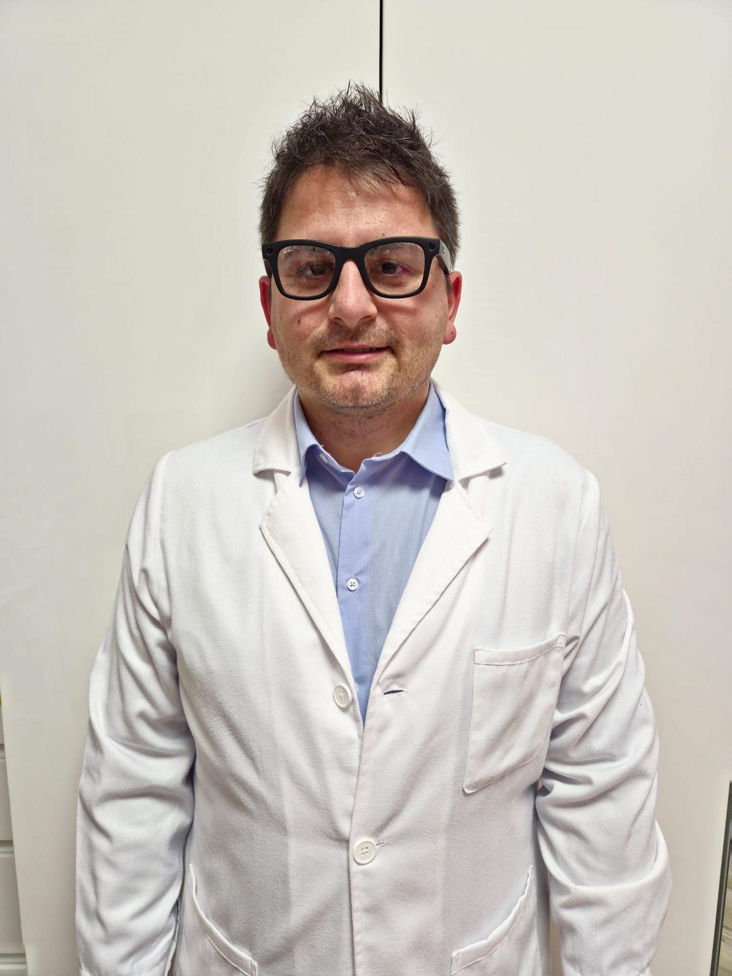 Man wearing glasses and a white lab coat standing indoors against a plain background.